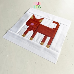 Könnte beinhalten: Ein weißer Stoffquadrat mit einem braunen und weißen Patchwork-Katzenmotiv. Die Katze hat eine rosa Nase und goldene Akzente.