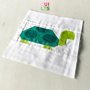 May include: A white fabric square with a green and white patchwork turtle design. The turtle is made of fabric squares sewn together. The fabric square has the text "QUILTS AND NICE STUFF" printed in the top center.