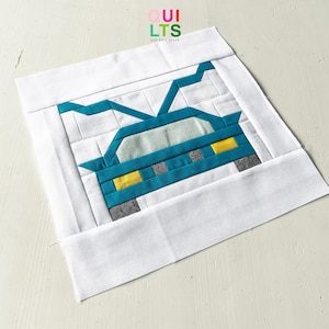 May include: A square fabric block featuring a teal car design with yellow headlights and gray tires, set against a white background. The block is framed with a white border. The text "QUILTS AND NICE STUFF" is in the upper right corner.
