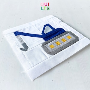 May include: A white fabric square with a blue and grey appliqué of an excavator. The excavator has yellow accents. The fabric square has a border of white fabric.