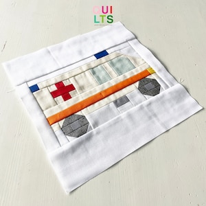 Könnte beinhalten: Ein handgefertigter Quiltblock mit einem Krankenwagen-Design. Der Krankenwagen besteht aus Stoffstücken in Weiß, Creme, Orange, Rot, Grau, Blau und Gelb. Der Block ist von einem weißen Rand umgeben. Der Text "QUILTS AND NICE STUFF" befindet sich in der oberen rechten Ecke.