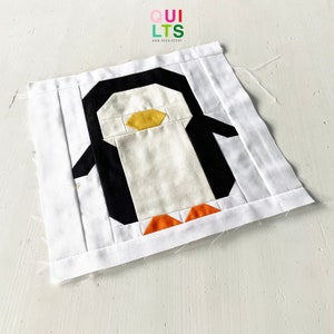 May include: A white fabric penguin quilt block with black, orange, and yellow fabric pieces. The block is on a white surface. "QUI LTS AND NICE STUFF" is written in colorful letters at the top of the image.