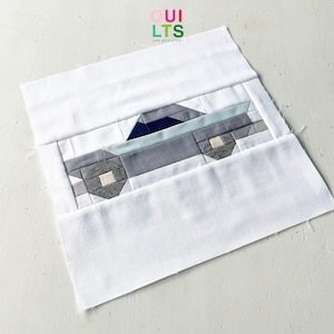 May include: A white fabric square with a blue, gray, and white patchwork car design in the center. The fabric square is folded in half, with the car design visible on the top half.