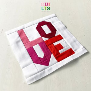 Könnte beinhalten: Ein weißer Stoffquadrat mit dem Wort "LOVE" in roten, rosa und weißen Stoffquadraten. Die Stoffquadrate sind zusammengenäht, um ein Patchwork-Design zu erstellen.