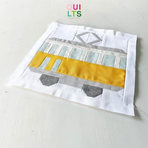 May include: A yellow and gray fabric patchwork quilt block depicting a vintage streetcar. The quilt block is on a white background. The text "QUI LTS AND NICE STUFF" is visible in the top left corner of the image.