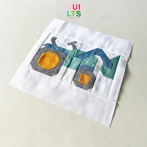 May include: A white fabric square with a patchwork design of a blue and green tractor. The tractor has a yellow circle for a wheel and a smaller yellow square for another wheel.