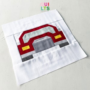 May include: A red, white, and gray patchwork quilt block featuring a car design. The car has yellow headlights and a gray front bumper.