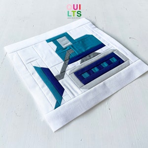 May include: A blue, grey and white fabric quilt block depicting a construction vehicle, a bulldozer. The quilt block is made using the paper piecing technique. The quilt block is on a white background.