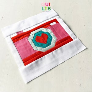 May include: A red, pink, and white fabric camera block with a teal heart in the center. The block is surrounded by a white border.