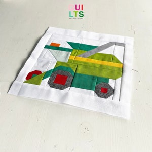 May include: A green and gray patchwork quilt block depicting a truck with red wheels and a yellow stripe. The block is surrounded by a white border. The text "QUI LTS AND NICE STUFF" is visible in the upper left corner of the image.