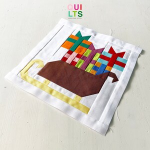 May include: A white fabric square with a brown, yellow, and multi-colored patchwork design of a sleigh filled with presents. The text "QUI LTS AND NICE STUFF" is printed in the top left corner.