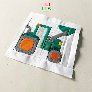 May include: A green and gray patchwork tractor quilt block with orange wheels. The block is made of fabric squares sewn together to create a geometric design. The quilt block is on a white background.