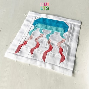 May include: A white quilt with a geometric pattern in shades of pink, red, blue, and green. The quilt features a jellyfish design with a blue bell and pink and red tentacles. The quilt is labeled "QUILTS AND NICE STUFF".