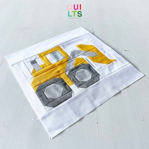 May include: A yellow and gray patchwork quilt block depicting a construction vehicle. The block is made of various fabric squares sewn together to create the image of a construction vehicle. The quilt block is on a white background.