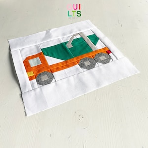 May include: A patchwork quilt block depicting a green and orange cement truck. The truck is made of various fabric squares and is surrounded by a white background.
