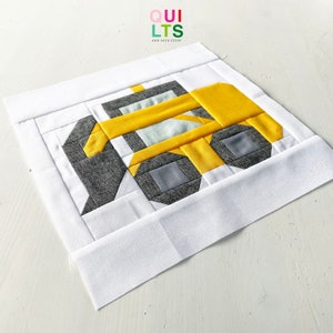 May include: A yellow and grey patchwork quilt block depicting a construction vehicle. The block is made of fabric squares sewn together to create the design. The quilt block is on a white background.