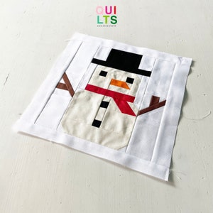 May include: A white fabric snowman quilt block with brown stick arms, a black top hat, a red scarf, and black and white buttons. The block is on a white surface.