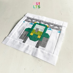 May include: A green, gray, and yellow patchwork quilt block featuring a geometric design of a truck. The block is surrounded by a white border.