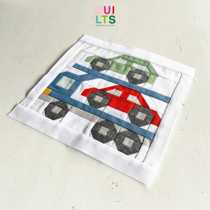 May include: A white fabric square with a blue, red, and green car quilt block. The quilt block features three cars stacked on top of each other. The top car is green, the middle car is red, and the bottom car is blue. The quilt block is surrounded by a white border.