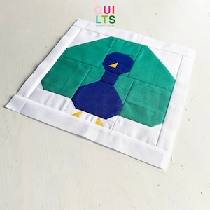 Könnte beinhalten: Ein blauer und grüner Patchwork-Quiltblock mit einem stilisierten Vogelmotiv. Der Vogel hat einen blauen Körper, gelbe Füße und einen gelben Schnabel. Der Quiltblock ist von einem weißen Rand umgeben.