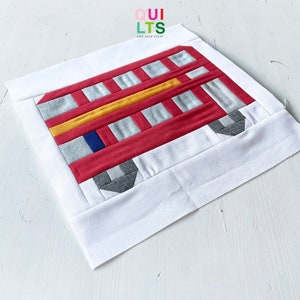 May include: A handmade quilt block depicting a red bus with silver, yellow, and blue details. The bus design is set against a white background and framed by a white border. The text "QUILTS AND NICE STUFF" is in the upper right corner.