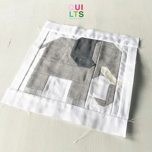 Può includere: Un blocco di trapunta a forma di elefante grigio e bianco con un bordo bianco. Il blocco è composto da diverse tonalità di grigio e bianco, e la proboscide dell'elefante è di una tonalità di grigio più chiara. Il blocco di trapunta è su una superficie bianca.