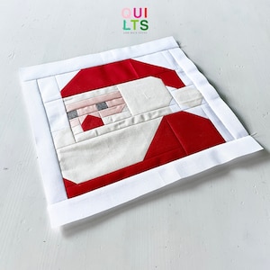 Könnte beinhalten: Ein weißer Stoffquadrat mit einem roten und weißen Patchwork-Design eines Weihnachtsmann-Gesichts. Der Stoffquadrat ist von einem weißen Rand umgeben. Der Text "QUI LTS AND NICE STUFF" ist in der oberen linken Ecke des Bildes gedruckt.