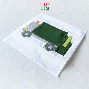 May include: A white fabric square with a green, gray, and yellow patchwork design in the center. The design is a simple geometric pattern that resembles a truck. The text "QUI LTS AND NICE STUFF" is printed in the top left corner.