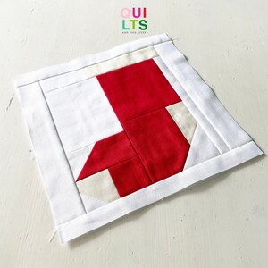 May include: A red and white fabric stocking block, with a white border, for a quilt. The block is made of squares and rectangles of fabric, sewn together to create the shape of a stocking. The fabric is a light cream color and a bright red.