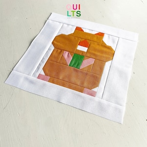 Könnte beinhalten: Ein brauner, rosa, grüner und orangefarbener Patchwork-Quiltblock mit einem stilisierten Tiermotiv. Der Block ist von einem weißen Rand umgeben.