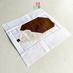 Könnte beinhalten: Ein weißer Stoffquadrat mit einer braunen und weißen Stoff-Igel-Applikation in der Mitte. Der Stoffquadrat hat einen weißen Stoffrand. Der Igel besteht aus zusammengenähten braunen und weißen Stoffstücken.