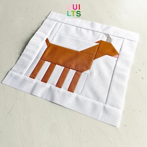 Könnte beinhalten: Ein brauner und weißer Patchwork-Quiltblock mit einem stilisierten Tierdesign. Das Tier ist eine braune Kuh mit weißen Streifen an den Beinen und einem weißen Gesicht. Der Quiltblock ist von einem weißen Rand umgeben.