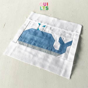 May include: A white fabric square with a blue whale applique. The whale is made of blue fabric with white and teal accents. The fabric square has a white border and the text "QUI LTS AND NICE STUFF" is printed in the top left corner.
