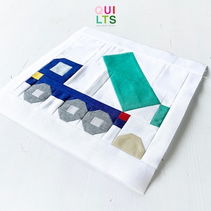 May include: A white fabric square with a blue truck and a green dump truck bed. The truck has gray wheels and a yellow detail. The truck is carrying a tan pile of dirt. The fabric square has the text "QUI LTS AND NICE STUFF" in rainbow colors.