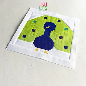 Könnte beinhalten: Ein grüner und blauer Patchwork-Quiltblock mit einem Pfauen-Design. Der Pfau hat einen blauen Körper und einen gelben Schnabel. Der Quiltblock ist von einem weißen Rand umgeben.