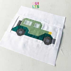 May include: A green and gray patchwork quilt block depicting a car. The car has a yellow headlight and a gray tire. The block is made of various shades of green and gray fabric.