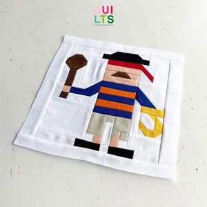 Könnte beinhalten: Ein farbenfroher Patchwork-Quiltblock mit einem Cartoon-Piraten, der eine Schatztruhe und einen Holzschläger hält. Der Pirat trägt ein rotes Bandana, ein blau-orange gestreiftes Hemd, eine beige Hose und schwarze Schuhe. Der Quiltblock ist aus verschiedenen Stoffen gefertigt, darunter Braun, Rot, Blau, Orange, Beige und Schwarz.