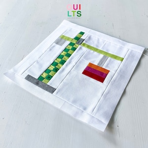 May include: A white fabric square with a green, gray, and orange geometric design. The design features a green and white checkered pattern, a gray and white striped pattern, and a rectangle of orange, pink, and red fabric.