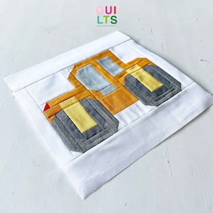 May include: A yellow and gray fabric patchwork quilt block depicting a construction vehicle. The block is on a white background. The text "QUI LTS AND NICE STUFF" is in the upper left corner of the image.