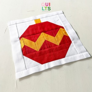 May include: A red and yellow patchwork Christmas ornament quilt block. The block is made of fabric squares sewn together in a zig-zag pattern. The block is surrounded by a white border.