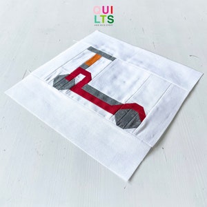 May include: A white fabric square with a red, gray, and orange geometric design. The design is a stylized letter L. The fabric square is on a white surface. The text "QUI LTS AND NICE STUFF" is in the upper left corner of the image.
