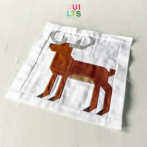 May include: A white fabric square with a brown and tan geometric deer design. The fabric square has a white border and the text "QUI LTS AND NICE STUFF" is printed in the top left corner.