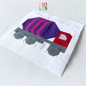 Könnte beinhalten: Ein weißer Stoffquadrat mit einem Patchwork-Design eines Zementlastwagens. Der LKW besteht aus violetten, rosa, roten und grauen Stoffstücken. Der LKW befindet sich auf einem weißen Hintergrund mit dem Text "QUILTS AND NICE STUFF" in der oberen linken Ecke.