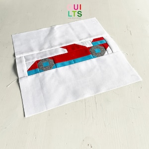 May include: A red, blue, and gray fabric race car applique on a white background. The applique is made up of several pieces of fabric sewn together.