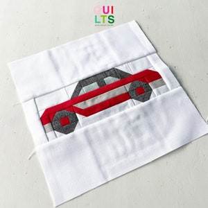 May include: A red and gray patchwork car quilt block. The car is made of various fabric squares and is outlined in white. The block is on a white background.