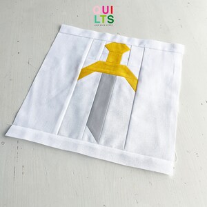 Könnte beinhalten: Ein weißer Stoffquadrat mit einem gelben und grauen Schwertmuster. Der Stoff ist gefaltet und hat einen weißen Rand.