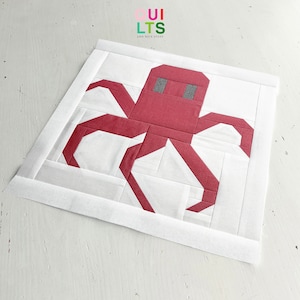 May include: A red and white quilt block featuring a stylised octopus design. The octopus is made up of red fabric squares and the background is white fabric. The quilt block is framed with a white border.