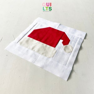 May include: A white fabric square with a red and white Santa hat applique. The fabric is folded over on all sides, creating a border around the applique.