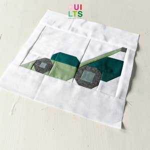 May include: A fabric quilt block featuring a green and gray tractor design. The tractor has two gray wheels, a green body, and a light green arm. The block is set against a white background. The text "QUILTS AND NICE STUFF" is in the upper right corner.