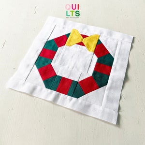 May include: A white fabric square with a green and red wreath design and a yellow bow. The wreath is made of fabric squares sewn together. The fabric square has the text "QUILTS AND NICE STUFF" in the top center.
